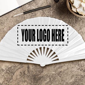 May include: A white folding fan with a semi-circular shape. The fan has a rectangular area with the text "YOUR LOGO HERE" in bold, black letters. The fan is lying on a textured, grey surface.