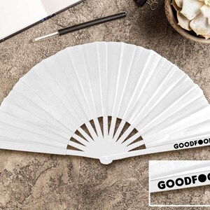 May include: A white paper fan, folded in a semi-circular shape. The fan features vertical pleats and a handle with the text "GOODFOOD" printed in black. The fan is open and displayed on a textured surface.