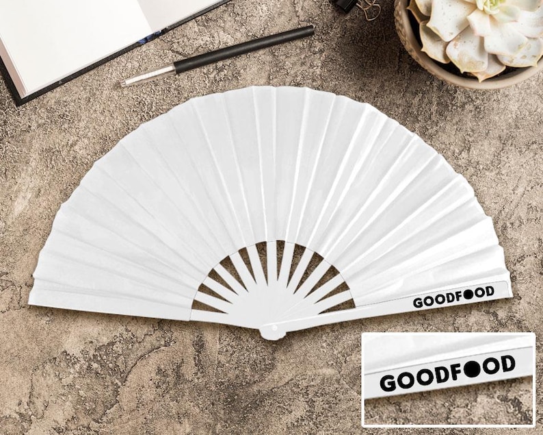 May include: A white paper fan, folded in a semi-circular shape. The fan features vertical pleats and a handle with the text "GOODFOOD" printed in black. The fan is open and displayed on a textured surface.