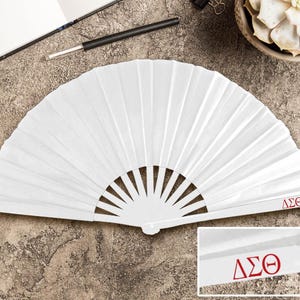 May include: A white, pleated paper fan with a semi-circular shape. The fan has a series of vertical folds and a handle. The letters "&Delta;&Sigma;&Theta;" are printed in red on the fan's handle. The fan is open and lying on a textured surface.