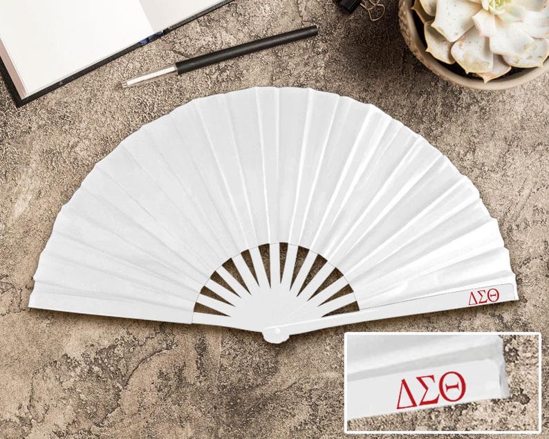 May include: A white, pleated paper fan with a semi-circular shape. The fan has a series of vertical folds and a handle. The letters "&Delta;&Sigma;&Theta;" are printed in red on the fan's handle. The fan is open and lying on a textured surface.