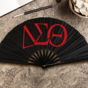 May include: A black folding fan with red Greek letters. The fan is open, displaying the letters Delta, Sigma, and Theta. The fan is on a textured, neutral-toned surface, with a pen, notebook, and succulent plant in the background.