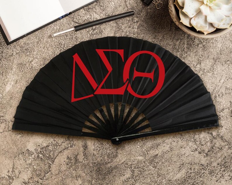May include: A black folding fan with red Greek letters. The fan is open, displaying the letters Delta, Sigma, and Theta. The fan is on a textured, neutral-toned surface, with a pen, notebook, and succulent plant in the background.