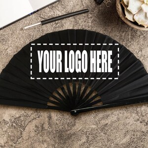 May include: A black folding fan with the text "YOUR LOGO HERE" in a white rectangle. The fan is open, showing its pleated design. The background is a textured, neutral surface.