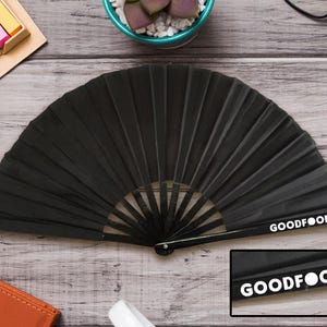 May include: A black folding fan with a ribbed design, lying open on a wooden surface. The text "GOODFOOD" is printed in white on the fan's handle. The fan is a dark, solid colour.
