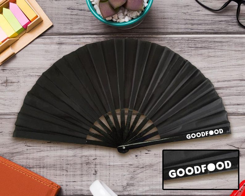 May include: A black folding fan with a ribbed design, lying open on a wooden surface. The text "GOODFOOD" is printed in white on the fan's handle. The fan is a dark, solid colour.