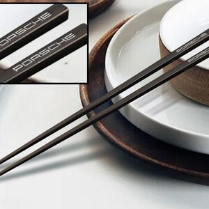 May include: A pair of black chopsticks with the word "PORSCHE" engraved on each chopstick. The chopsticks are on a white plate with a brown rim.