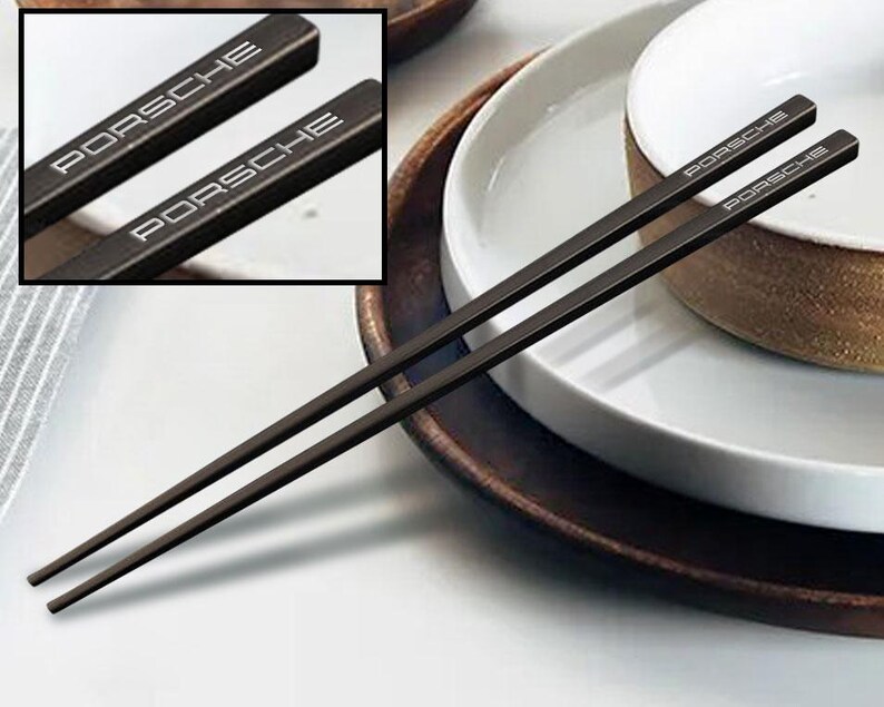 May include: A pair of black chopsticks with the word "PORSCHE" engraved on each chopstick. The chopsticks are on a white plate with a brown rim.
