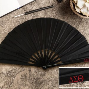May include: A black pleated folding fan, featuring a red embroidered "&Delta;&Sigma;&Theta;" emblem. The fan is open and displayed on a textured surface. A pen and notebook are visible in the background.