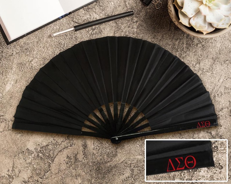 May include: A black pleated folding fan, featuring a red embroidered "&Delta;&Sigma;&Theta;" emblem. The fan is open and displayed on a textured surface. A pen and notebook are visible in the background.