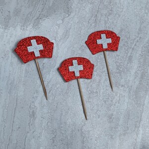 May include: Three red and white glitter nurse hat cupcake toppers with white crosses. The toppers are on wooden sticks.