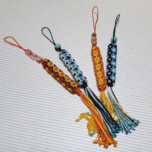 May include: Four handmade macrame keychains with colorful tassels. The keychains feature intricate woven patterns in shades of orange, yellow, blue, and white, with small beads and loops for attachment. Each keychain is unique.