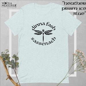 May include: A light blue t-shirt featuring a black dragonfly graphic and the text "dinna fash sassenach". The text is in a circular design around the dragonfly. The shirt is a heather prism ice blue colour.