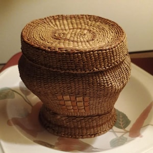 May include: A woven basket with a lid. The basket is made of natural-colored fibers, with a round lid and a bulbous body. The basket sits on a decorative plate with floral designs.