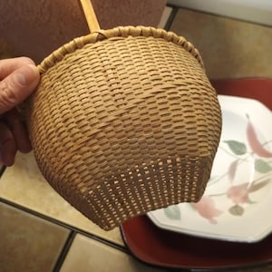 May include: A light tan woven basket with a square base that transitions into a rounded top rim. The basket features a single vertical wooden handle extending upward from the center of the rim, constructed from thin, flexible strips of natural material.