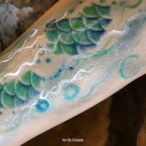 May include: A close-up of a mermaid tail painted on a person's arm. The tail is green and blue with white scales. The background is a silver glitter with white swirls and blue circles.