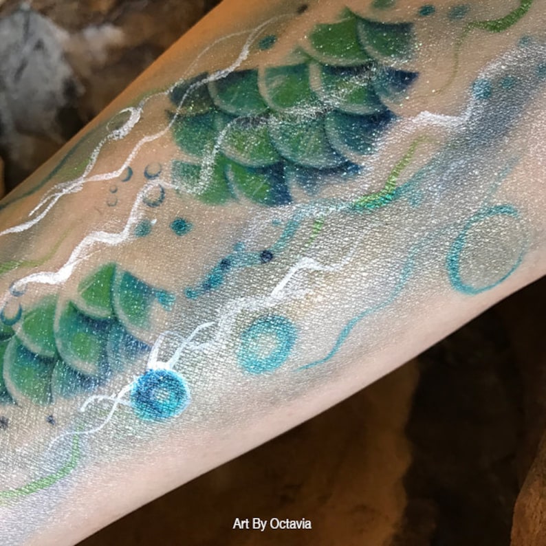 May include: A close-up of a mermaid tail painted on a person's arm. The tail is green and blue with white scales. The background is a silver glitter with white swirls and blue circles.
