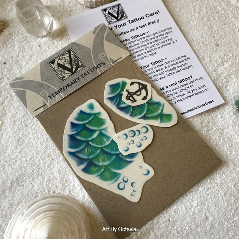May include: Two temporary tattoos on a brown card. One tattoo is a mermaid tail with green and blue scales. The other tattoo is an anchor with a rope. The card has the text "Temporary Tattoos" and "Art By Octavia".