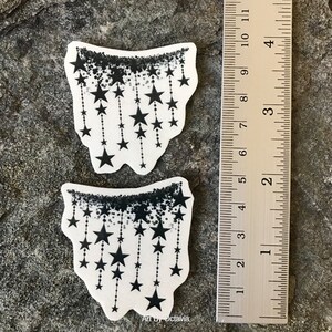 K&ouml;nnte beinhalten: Zwei wei&szlig;e Aufkleber mit einem schwarzen Sterndesign. Jeder Aufkleber hat einen schwarzen, gesprenkelten Rand oben, mit schwarzen Sternen, die an gepunkteten Linien herunterh&auml;ngen. Ein Lineal befindet sich neben den Aufklebern und zeigt Zentimeter.