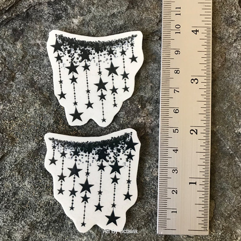 K&ouml;nnte beinhalten: Zwei wei&szlig;e Aufkleber mit einem schwarzen Sterndesign. Jeder Aufkleber hat einen schwarzen, gesprenkelten Rand oben, mit schwarzen Sternen, die an gepunkteten Linien herunterh&auml;ngen. Ein Lineal befindet sich neben den Aufklebern und zeigt Zentimeter.