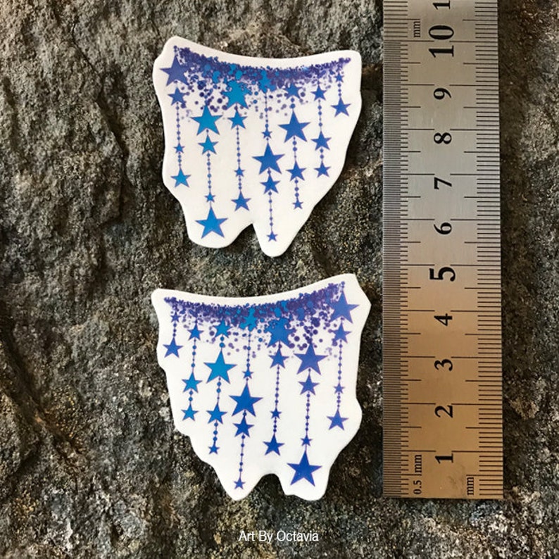 K&ouml;nnte beinhalten: Zwei wei&szlig;e Aufkleber mit blauem Sternmuster. Die Aufkleber haben einen gezackten Rand und eine blaue Glitzeroberfl&auml;che mit h&auml;ngenden blauen Sternen. Der Text "Art By Octavia" befindet sich unten. Ein Lineal zeigt, dass die Aufkleber etwa 5 cm lang sind.