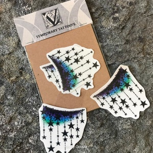 K&ouml;nnte beinhalten: Bl&auml;tter mit tempor&auml;ren Tattoos mit einem Sterndesign. Die Tattoos haben einen Farbverlauf aus Blau, Lila und Schwarz, mit schwarzen Sternen, die an gepunkteten Linien h&auml;ngen. Die Verpackung tr&auml;gt die Aufschrift "TEMPORARY TATTOO'S" und "Arty Octavia".