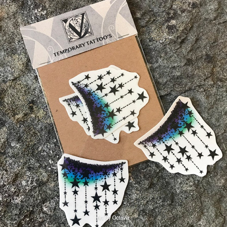 K&ouml;nnte beinhalten: Bl&auml;tter mit tempor&auml;ren Tattoos mit einem Sterndesign. Die Tattoos haben einen Farbverlauf aus Blau, Lila und Schwarz, mit schwarzen Sternen, die an gepunkteten Linien h&auml;ngen. Die Verpackung tr&auml;gt die Aufschrift "TEMPORARY TATTOO'S" und "Arty Octavia".