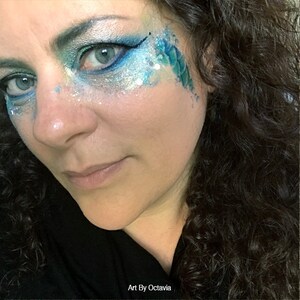 May include: A woman with dark curly hair has blue and silver glitter makeup on her face. The makeup includes a blue and green scale design on her cheek. The text "Art By Octavia" is at the bottom of the image.
