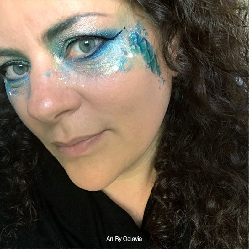 May include: A woman with dark curly hair has blue and silver glitter makeup on her face. The makeup includes a blue and green scale design on her cheek. The text "Art By Octavia" is at the bottom of the image.