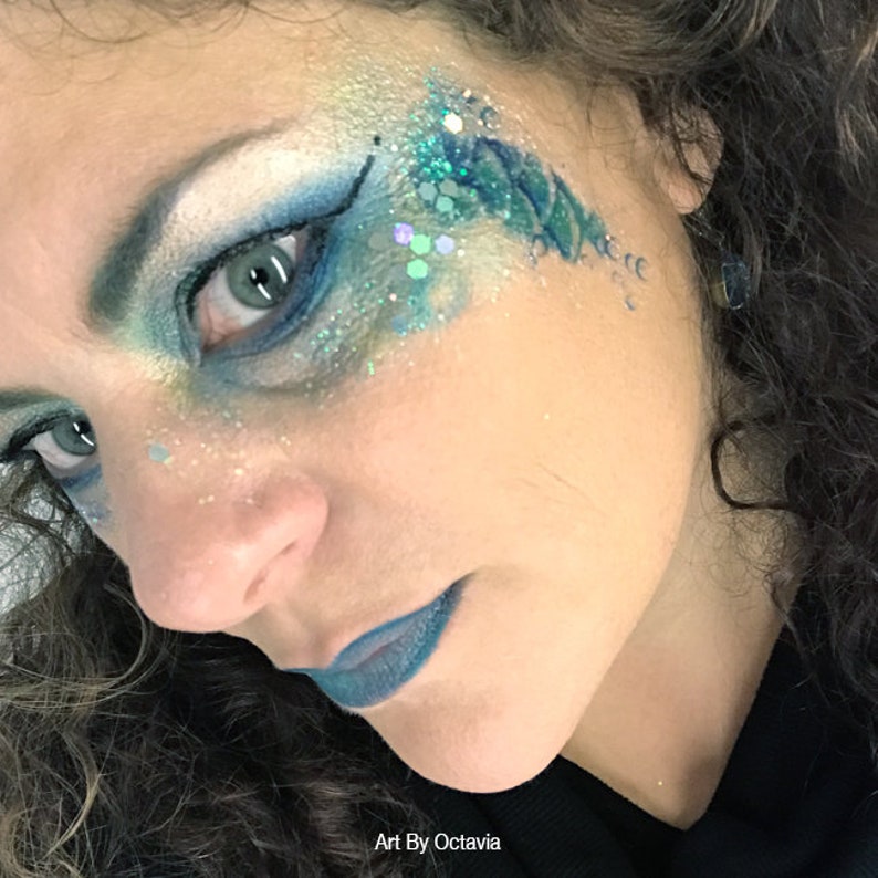 May include: A close-up of a person's face with blue and green glitter makeup applied around the eye. The makeup is designed to look like a mermaid's tail. The text "Art By Octavia" is at the bottom of the image.