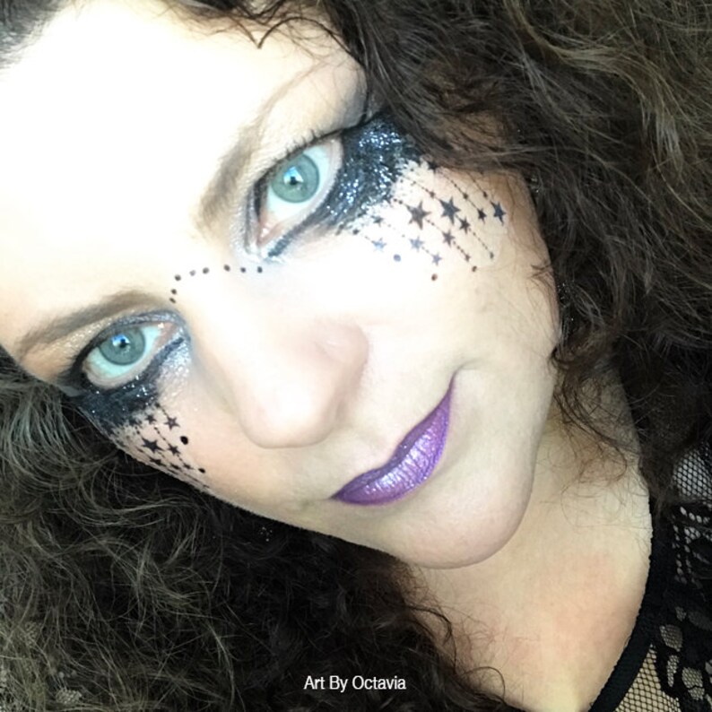 K&ouml;nnte beinhalten: Nahaufnahme eines Gesichts mit dramatischem Augen-Make-up und lila Lippenstift. Das Augen-Make-up zeigt schwarzen Glitzer und Sternakzente. Der Text "Art By Octavia" ist unten links sichtbar.