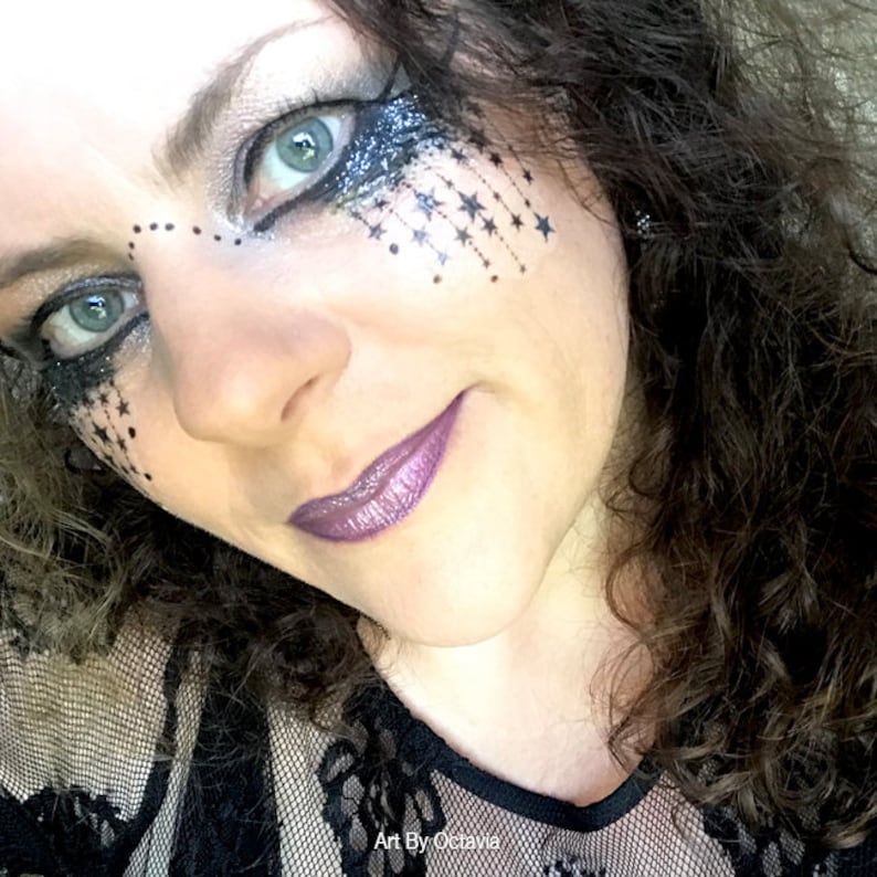 K&ouml;nnte beinhalten: Nahaufnahme einer Person mit aufwendigem Augen-Make-up mit silbernen Glitzer- und schwarzen Sternakzenten. Die Person hat lila Lippenstift und dunkles, lockiges Haar. Das Bild ist mit "Art By Octavia" signiert.