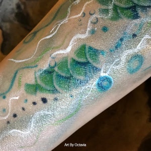 May include: A close-up of a person's arm with a blue, green, and white painted design. The design features waves, bubbles, and scales. The text "Art By Octavia" is visible at the bottom of the image.