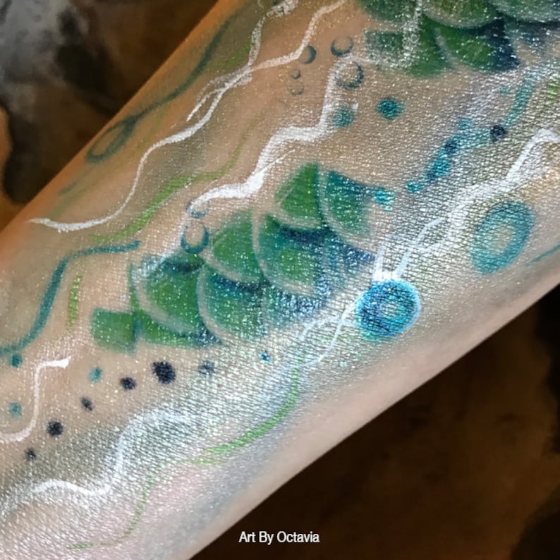 May include: A close-up of a person's arm with a blue, green, and white painted design. The design features waves, bubbles, and scales. The text "Art By Octavia" is visible at the bottom of the image.