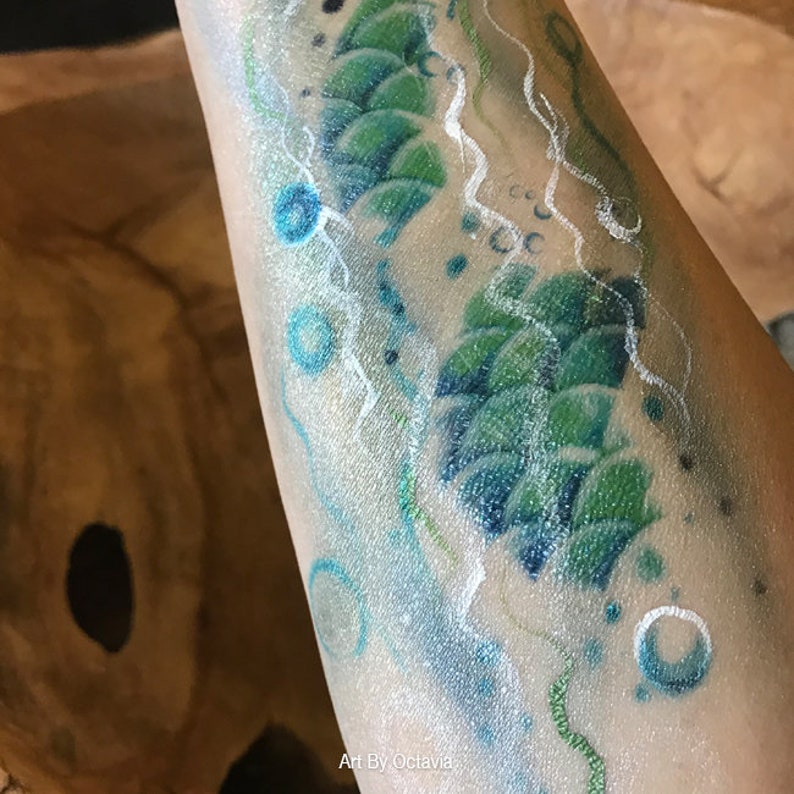 May include: A close-up of a green and blue mermaid tail painted on a person's arm. The tail is surrounded by white swirls and blue bubbles.