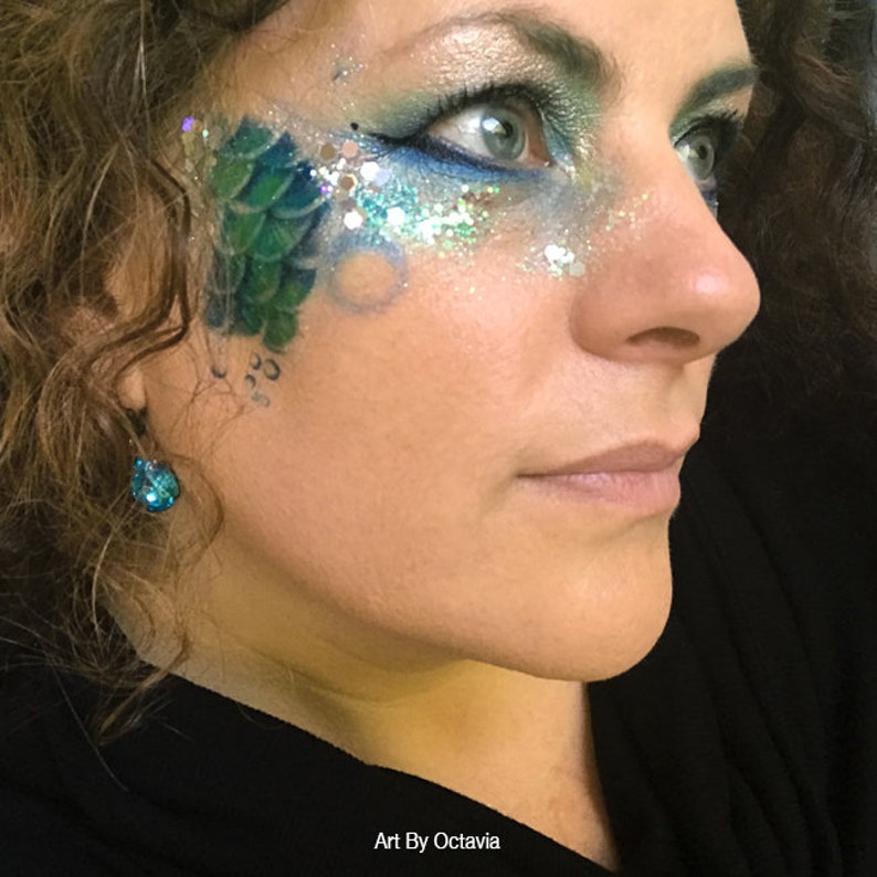 May include: A woman's face with blue and green glitter makeup. The makeup includes a green scale design on the cheek and blue glitter around the eye. The woman is wearing a blue gemstone earring.