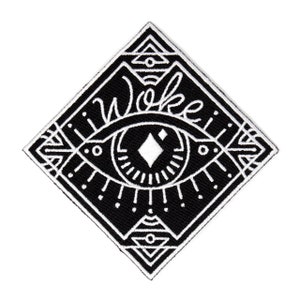 Op de afbeelding: Zwart-witte geborduurde patch met een gestileerd oog en geometrische vormen. Het woord "Woke" staat boven het oog.