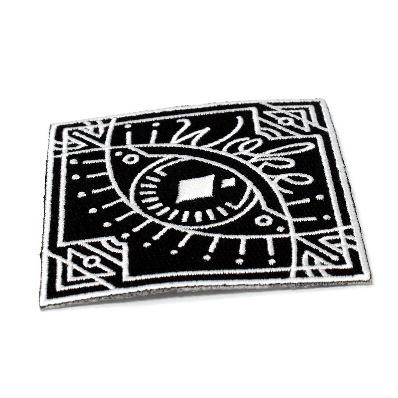 May include: Black and white embroidered patch with a stylized eye design. The eye is surrounded by geometric shapes and lines.