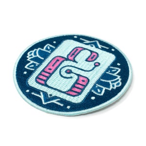 May include: A blue and pink embroidered patch featuring a stylised letter 'G' within a square. The patch is encircled by a dark blue circle with an ornate border.