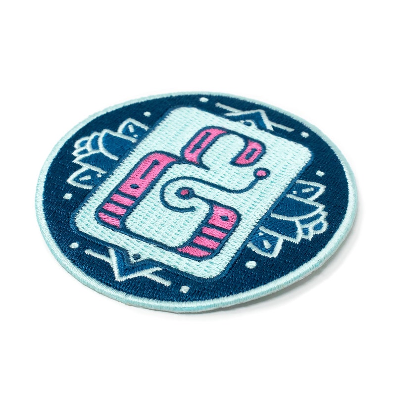 May include: A blue and pink embroidered patch featuring a stylised letter 'G' within a square. The patch is encircled by a dark blue circle with an ornate border.