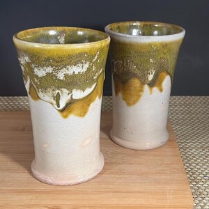 May include: Two handmade ceramic tumblers. The top rims are glazed in a mustard yellow, transitioning to a moss green, and then a creamy white. The lower portion of the tumblers is a soft, off-white color. The tumblers are displayed on a wooden cutting board.
