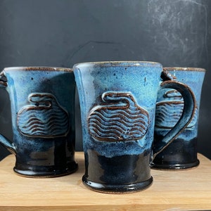 May include: Three blue and brown ceramic mugs with a curling stone design. The mugs have a dark blue glaze and a brown curling stone design.