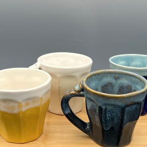 May include: Four ceramic mugs in shades of yellow, white, blue, and dark blue sit on a wooden tray. Each mug has a unique design and handle. The mugs are handmade and have a rustic, artisanal feel.