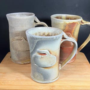May include: Three handmade ceramic mugs with unique designs. The mugs feature earthy tones of beige, brown, and blue-grey, with textured surfaces and distinctive handles. Each mug has a carved, wave-like design on the side, adding to their artisanal appeal.