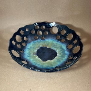 May include: A round ceramic bowl with a scalloped edge and a series of circular cutouts along the rim. The interior features a dark center that transitions into a vibrant green and blue gradient, while the exterior rim is a glossy black finish.