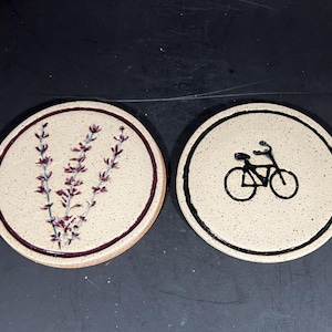 May include: Two round ceramic coasters. One features a lavender sprig design in purple and green. The other has a black bicycle graphic. Both have a speckled, off-white background and a dark brown border.