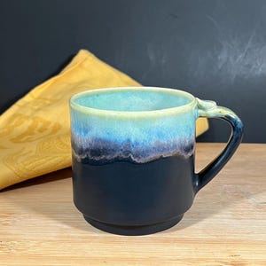May include: A ceramic mug with a gradient design. The top rim is light blue, transitioning to dark blue and black at the base. The handle is black. The mug sits on a wooden surface, with a yellow cloth in the background.
