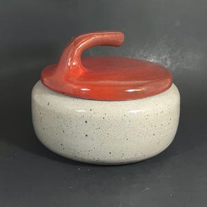 May include: A ceramic curling stone-shaped container with a red handle and lid. The container's body is speckled gray, and the lid and handle are a glossy red. The design is reminiscent of a curling stone used in the sport of curling.