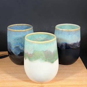 May include: Three ceramic stemless wine glasses with a unique color gradient. The glasses feature a white base, transitioning to green, then blue, and finally a dark blue or black rim. The glasses are displayed on a wooden surface.