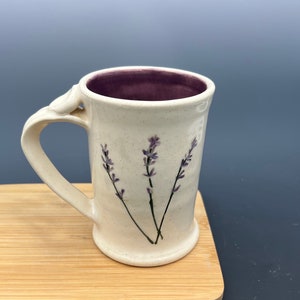 May include: White ceramic mug with a purple interior and a lavender floral design. The mug has a handle that is shaped like a leaf.